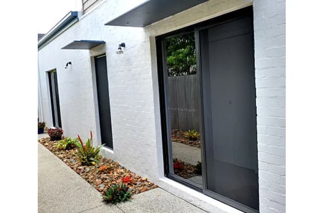 Dark security screens on the sliding door and windows of a white painted brick home with a tropical garden bed